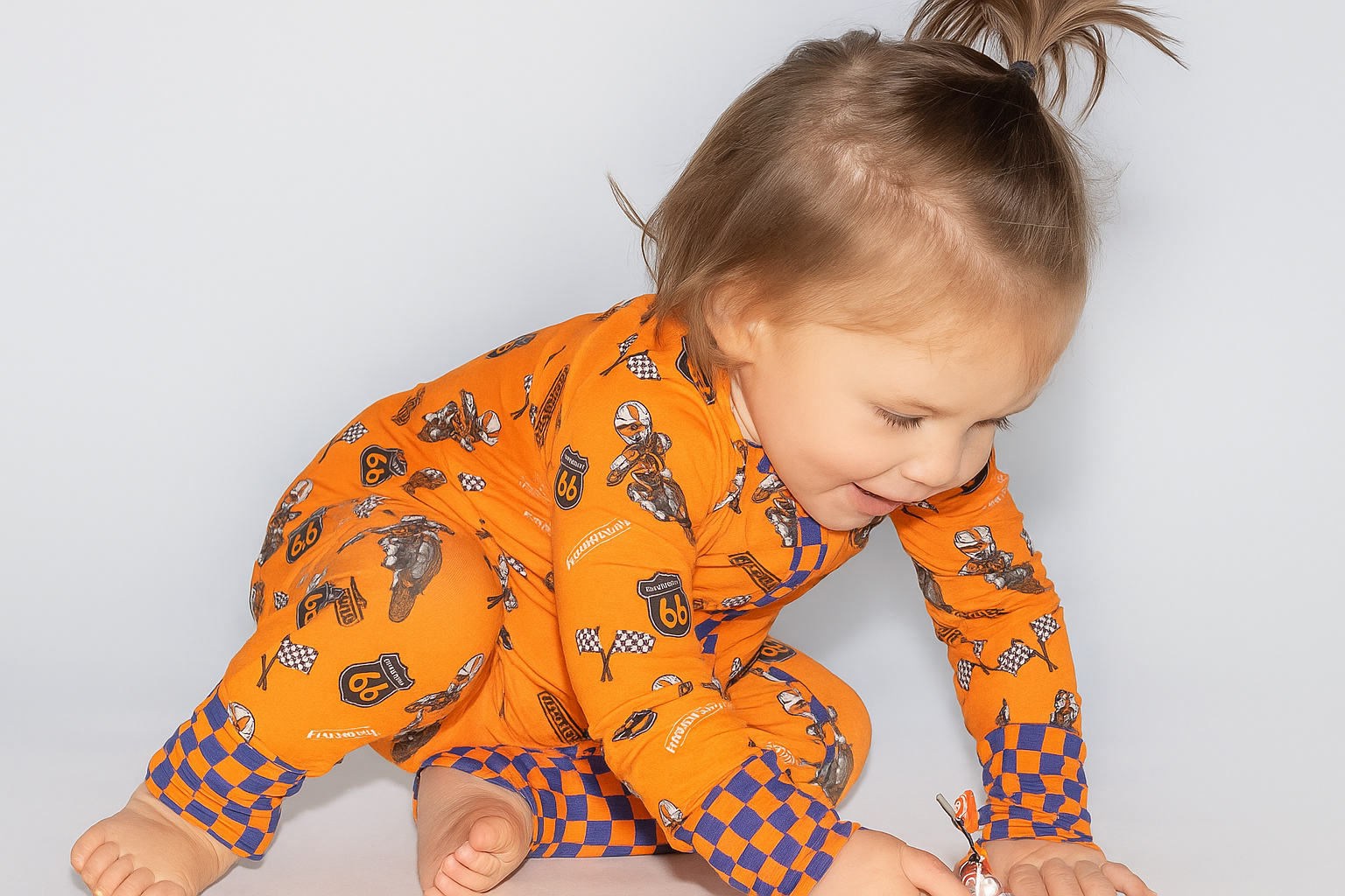 Baby in bright orange bamboo pajamas with racing and checker print, playing on the floor
