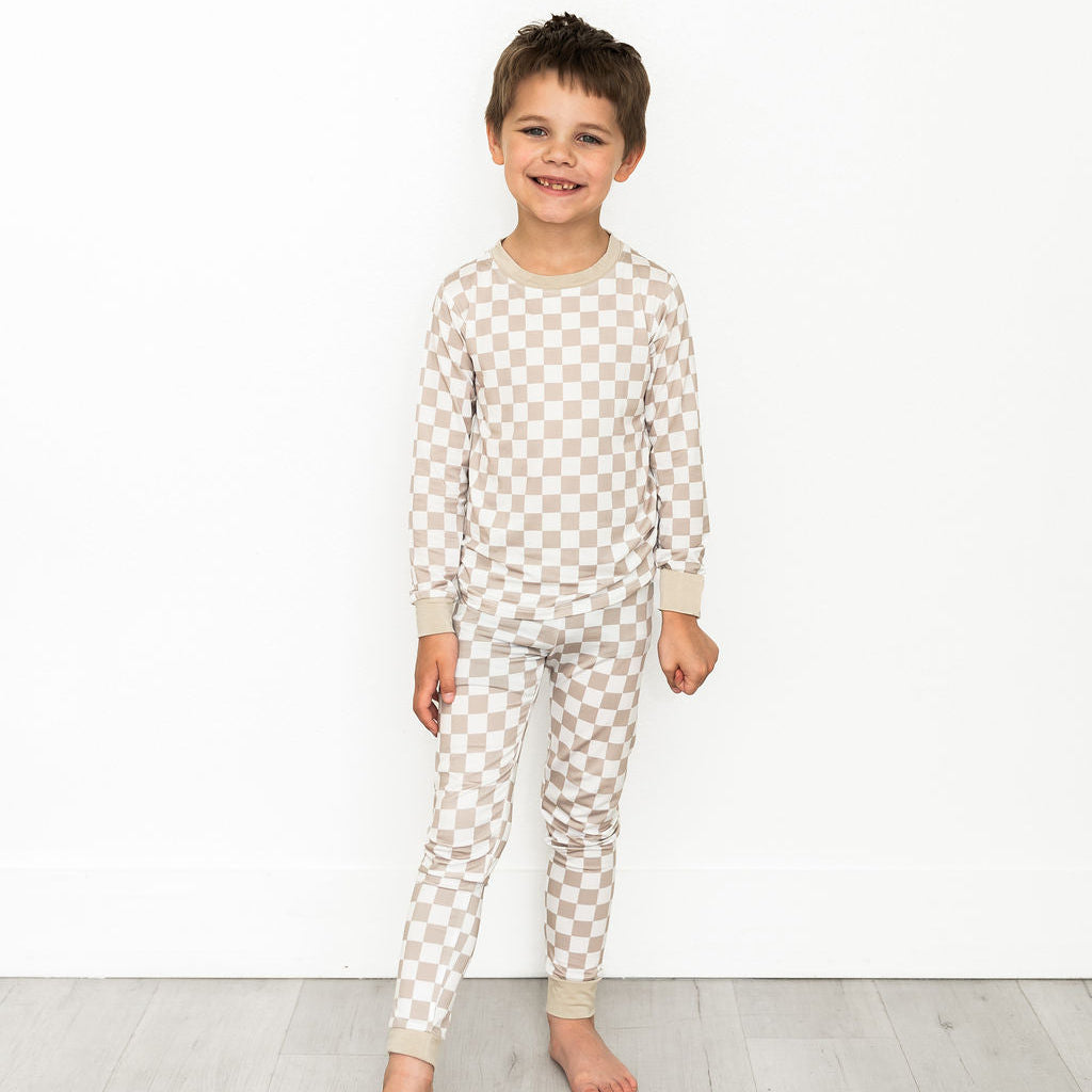 A young boy stands barefoot against a white wall and light wood floor, wearing a matching long-sleeve pajama top and pants set in a beige and white checkerboard pattern.