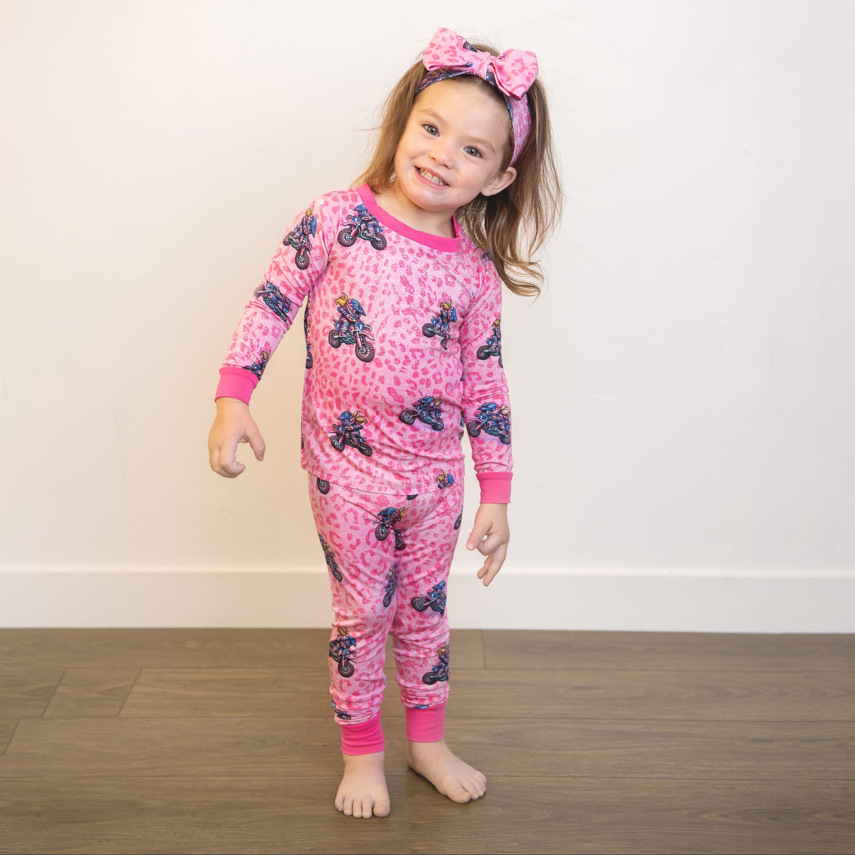 Child wearing pink pajamas with a pattern, standing on a wooden floor against a white wall.