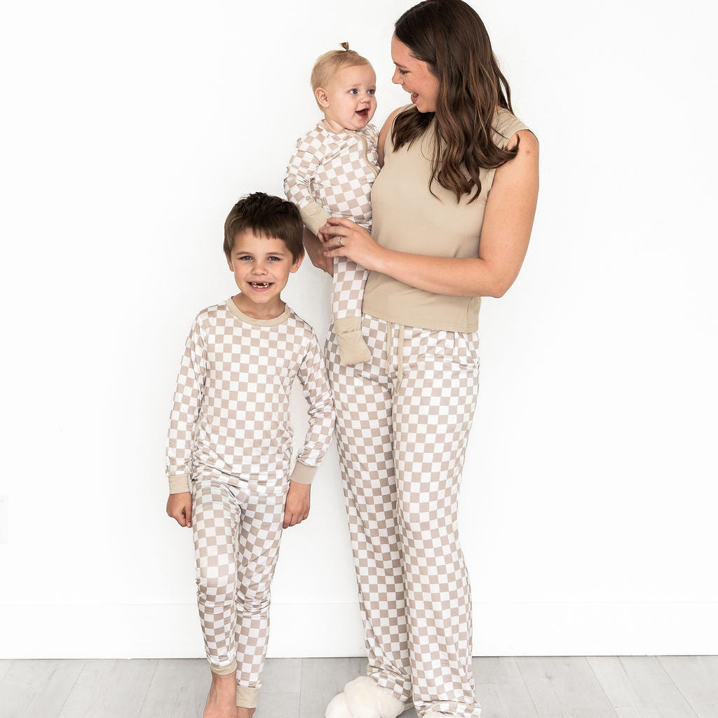 A woman and two young boys wearing matching beige and white checkered pajamas pose together against a clean white wall and light wood floor.