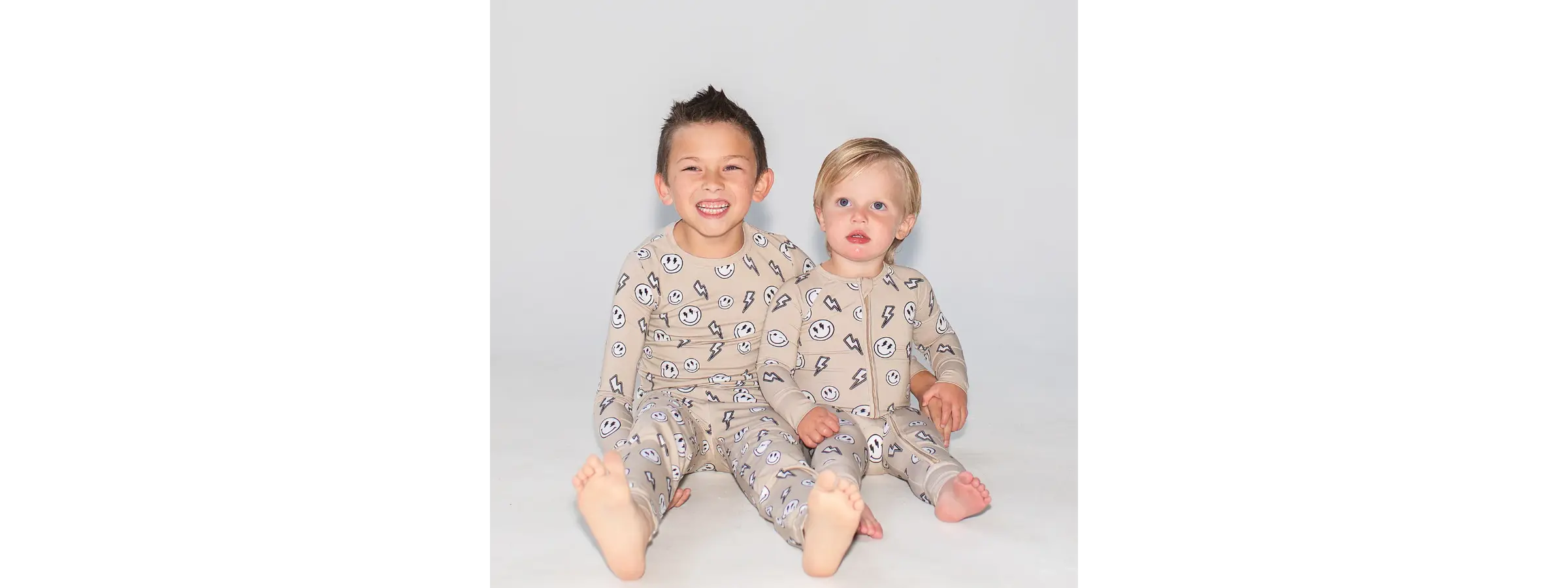 Two toddlers wearing beige convertible Khlobe Zippy pajamas with smiley and lightning print
