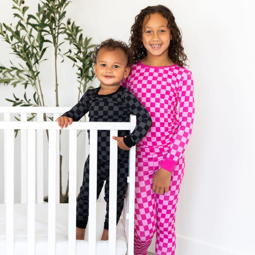 Two children in checkered pajamas standing next to a white crib with a plant in the background.