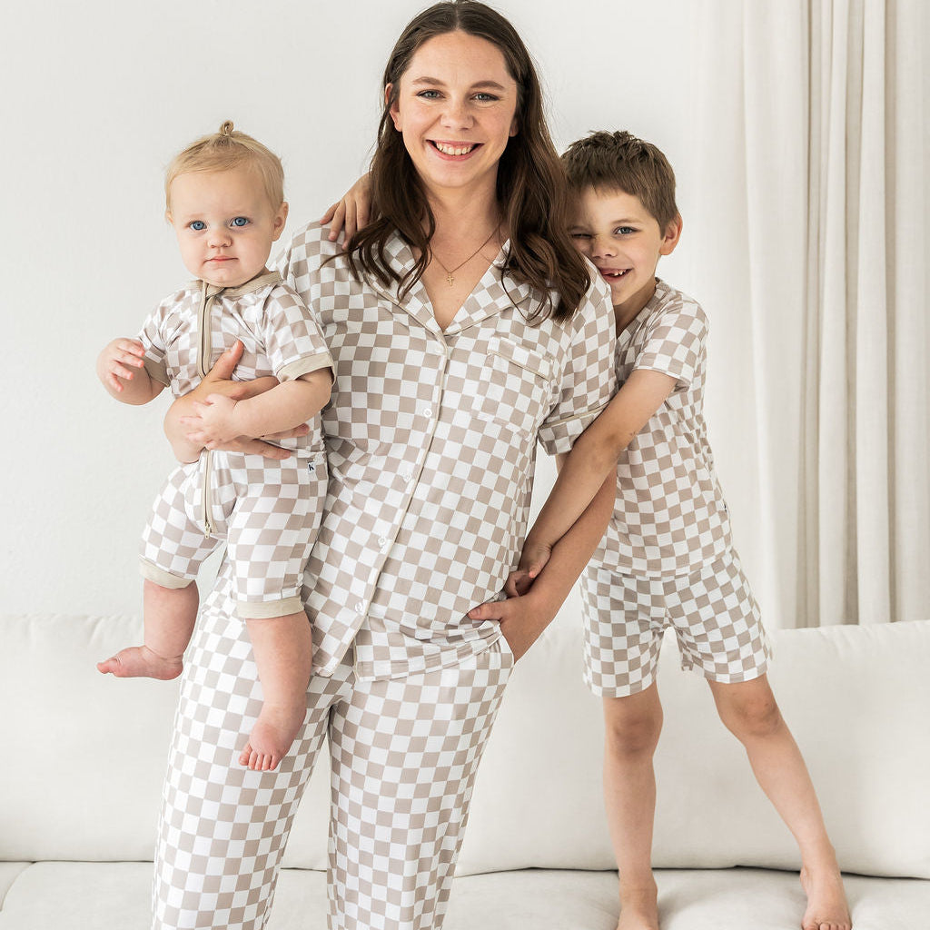 A woman and two children wearing matching short-sleeve pajamas with a beige and white checkerboard pattern, posed together on a white sofa in a bright room.