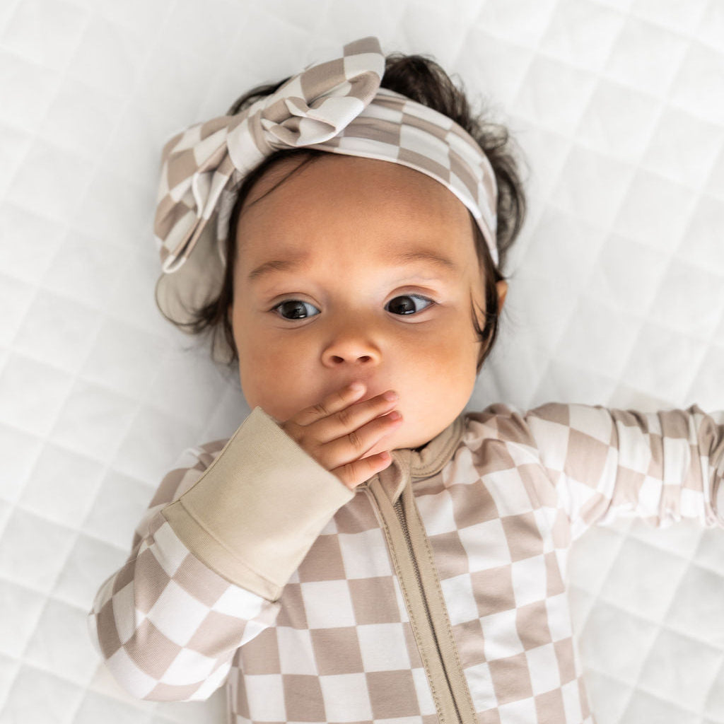 A baby lying on a white quilted mattress, wearing a beige and white checkered zip-up onesie and a matching checkered bow headband.