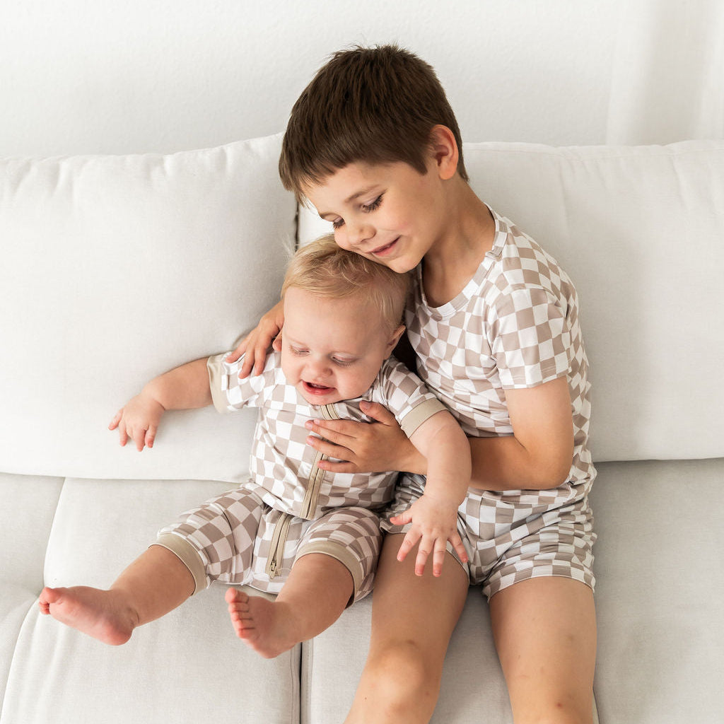 Two children wearing matching beige and white checkered short-sleeve tops and shorts sit together on a white couch. The older child is holding the younger child, who appears to be crying.