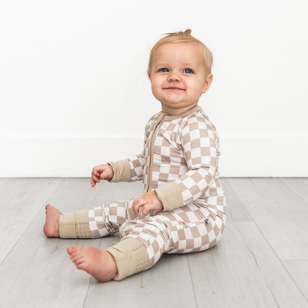 A baby sitting on a light gray wooden floor against a white wall, wearing a beige and white checkered one-piece pajama with beige cuffs.