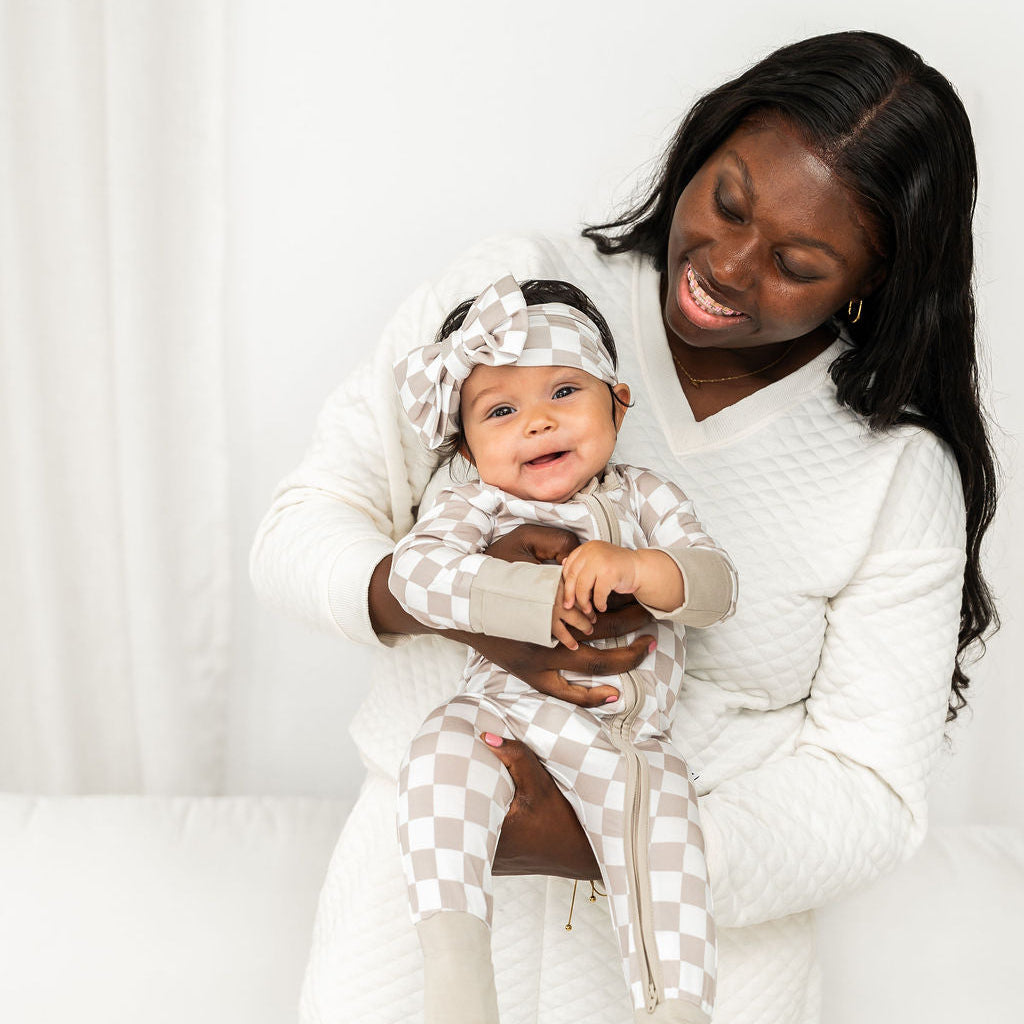 A smiling woman in a white quilted robe holds a baby wearing a matching white and beige checkered outfit with a checkered headband, set against a bright white background.